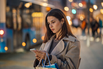 Young woman browsing on smartphone while holding shopping bags in city at night