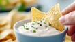 © Pinklife - A creamy white dip garnished with green parsley is shown in a blue bowl, highlighting the savory delight of enjoying snacks with crispy tortilla chips.