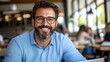 © Pinklife - A smiling man wearing glasses in a blue shirt is seated in a relaxed workspace, suggesting creativity and openness, with blurred people in the background working.