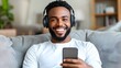© tuiphotoengineer - Cheerful African American Man Relaxing on Couch and Laughing While Using Smartphone and Wireless Headphones in Pastel Colored Modern Living Room Environment