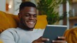 © tuiphotoengineer - Close up of a happy and smiling Black man using a tablet device while sitting comfortably on the sofa at home engrossed in a digital experience with a cybernetic and triadic color scheme