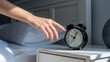 © ColorfulFlowerStudio - Person's hand reaching for alarm clock on bedside table in morning light.