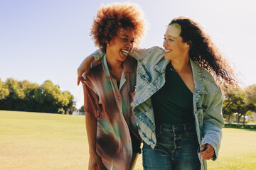 Two young girlfriends bonding outdoors, laughing and embracing on a sunny day in a green park landscape