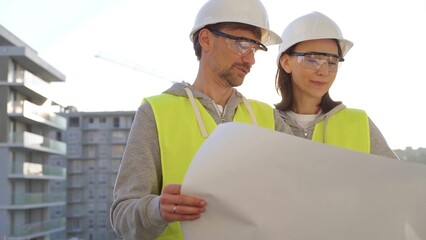 architects or engineers wearing safety helmets and vests holding blueprint and discussing something 