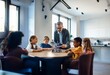 © YiuCheung - A teacher in a suit explains a robotics project to a diverse group of students sitting around a table.