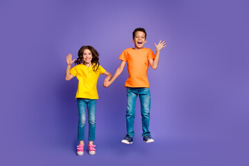 Wall Mural - Full length photo of lovely two little kids jump wave hand dressed stylish orange yellow garment isolated on purple color background