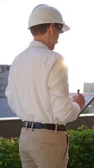 Man a construction engineer wearing white shirt and hard hat is making notes on a clipboard while inspecting a building site at sunset, back vertical view . Architecture concept