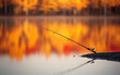 © Suritong - Fishing rod resting on the shore, calm water reflecting autumn colors.