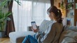 © COK House - A woman in a gray sweatshirt sits on a couch in a modern living room, using a phone app. The setting is casual and relaxed, highlighting home comfort.