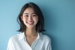 © Surachetsh - Beautiful Asian girl with long dark hair stands smiling warmly in front of a turquoise background. She is wearing a simple white shirt, exuding natural beauty and positivity.