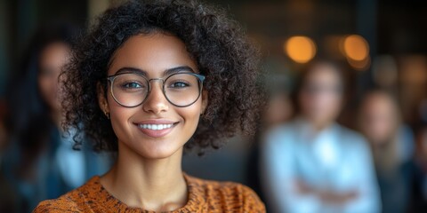 A young woman with curly hair and glasses smiling confidently in a blurred background setting, possibly depicting a professional or social environment