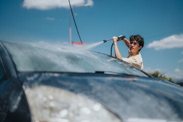 A young man uses a high-pressure hose to wash his car on a sunny day. The vibrant scene captures the essence of summer chores and outdoor maintenance.