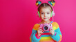 © Curioso.Photography - Young girl in colorful rainbow sweater holding a pink camera against a bright pink background, showcasing creativity and childhood fun.