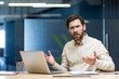 © Tetiana - Portrait of an upset and disappointed young businessman sitting in the office at a desk with a laptop, looking at the camera and spreading his hands