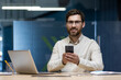 © Tetiana - Close-up portrait of a young smiling man in glasses and a shirt sitting at a desk in the office, holding a mobile phone and looking at the camera