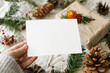 © sonyachny - Christmas card mock up. Hand holding empty greeting card on rustic table with fir branches and pine cones. Space for text. Season's greetings. Zero waste postcard template. Merry Christmas