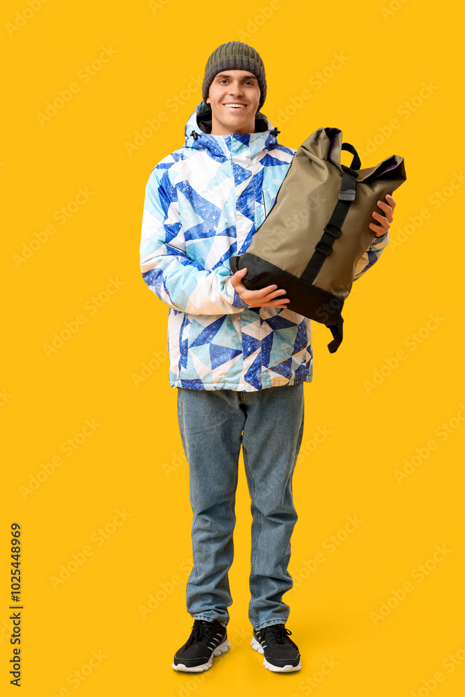 Young man in stylish puffer jacket and hat with bag on yellow background