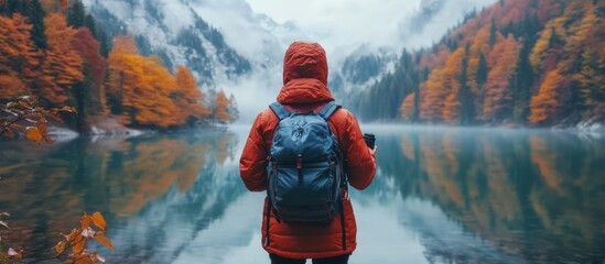 Wall Mural - a lone hiker in a red jacket stands by a misty lake surrounded by autumn foliage, looking out at the