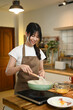 © Prathankarnpap - Cheerful young asian woman cooking scrambled eggs in frying pan on a stovetop