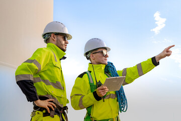 two engineers in high visibility gear and hard hats observe a wind turbine and communicate via radio