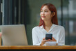 © Songsak C - Portrait of a beautiful confident businesswoman using a laptop computer holding a mobile phone sitting in a modern office. Smiling Asian freelancer working online from home.