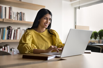 Cheerful successful young Indian project manager woman enjoying online communication at computer, typing on laptop in co-working space, smiling, working, using job application, browsing Internet