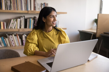 Wall Mural - Happy thoughtful young Indian professional woman working on research study in library, using laptop at table, looking away, smiling, thinking on successful business project solution