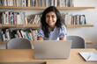 © fizkes - Happy beautiful Indian student woman working at laptop in library, posing for portrait at workplace table, looking at camera, bending over desktop, smiling, laughing