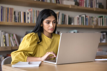 serious indian college student girl working at computer in public library, typing on laptop at table