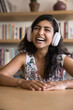 © fizkes - Cheerful young Indian student blogger girl in headphones looking at camera with toothy smile, laughing, talking on video call from library, posing for vertical portrait with bookshelves behind