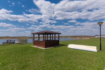 Wooden square gazebo on a green lawn, on the shore of a pond, against a beautiful blue .sky with clouds.