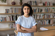 © fizkes - Positive confident Indian entrepreneur woman standing at workplace in co-working with library, posing with arms folded, looking at camera for business professional head shot portrait