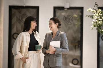 Canvas Print - Cheerful middle aged business woman talking to young colleague on office coffee break, walking, standing on hallway, speaking to coworker woman, laughing. Businesswomen discussing project