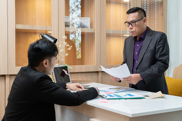 A man in a suit is pointing at a piece of paper on a desk. The man is wearing glasses and he is giving instructions or directions to the other person