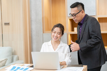 A man and a woman are sitting at a desk with a laptop. The man is pointing at the laptop screen