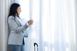 © roberto - Happy young business executive woman with suitcase traveling in a new city, removing the curtain from the hotel room window, looking out the window at the city views, business trips and work, resting