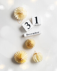 Flat lay of decorative ornaments and wood calendar date December 31st New Year's Eve, Minimal layout with paper festive decor beige balls, Christmas baubles on white wth bokeh, modern decor