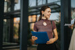 © qunica.com - Confident young woman smiling while holding documents outside an office building, showcasing a positive professional atmosphere during a business meeting.