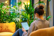 © alisluch - Woman sitting indoors, working on a tablet, surrounded by lush green plants, cozy yellow couch in background