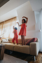 Wall Mural - Two happy girls in red dresses enjoying a playful dance on the sofa in a sunlit room. Capturing joy, fun, and carefree childhood moments indoors.