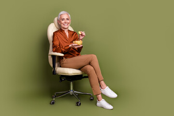 Poster - Full length photo of pensioner lady sit office armchair eat chips dressed brown formalwear isolated on khaki background