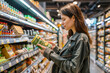 © Koshiro K - Young woman reading product label while shopping in a grocery store, carefully choosing packaged food items.