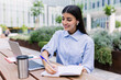 © Xavier Lorenzo - Young adult student woman taking notes while using laptop computer sitting outside of college building. Millennial female in smart casual clothes listening virtual video call. Business and education.