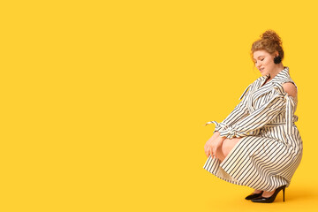 Wall Mural - Beautiful young woman in stylish striped dress sitting against yellow background