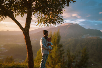 A man is gently holding a small baby in his arms while standing underneath a large, leafy tree that provides a cool, inviting shade