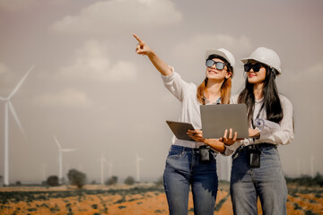 Two female engineer working outdoor with safety at wind turbines clean energy power station background, worker people with renewable energy technology for future concept.