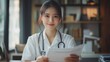 © ifoto - A Chinese female doctor wearing medical white and a stethoscope around her neck with a smile, sits at an office desk facing forward while holding paper documents in front of her