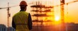 © Cheewynn - Silhouette of a construction worker standing on a building site at sunset, industrial scaffolding in the background, career in architecture and engineering