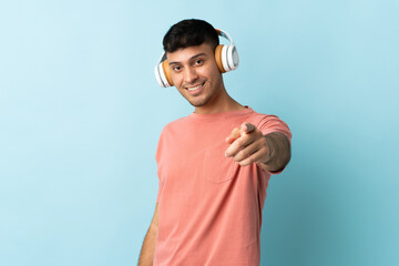 Young Colombian man isolated on blue background listening music and pointing to the front