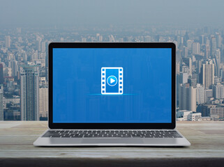 Play button with movie flat icon on modern laptop computer monitor screen on wooden table over city tower and skyscraper, Business cinema online concept